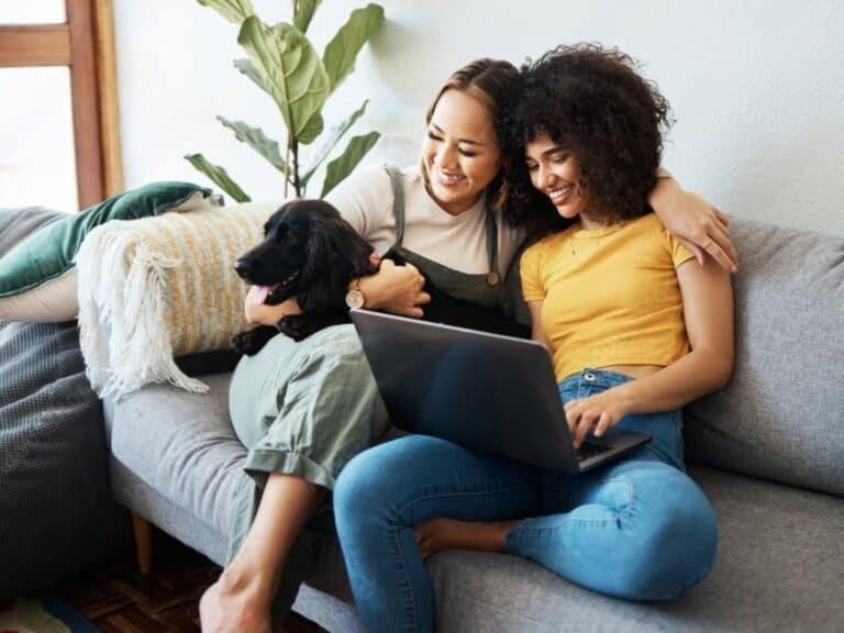 Two people sitting on a couch, smiling and embracing a black dog while one works on a laptop. The scene, perfect for a house sitter profile example, includes a vibrant potted plant in the background, illustrating how to write an inviting house sitter profile.