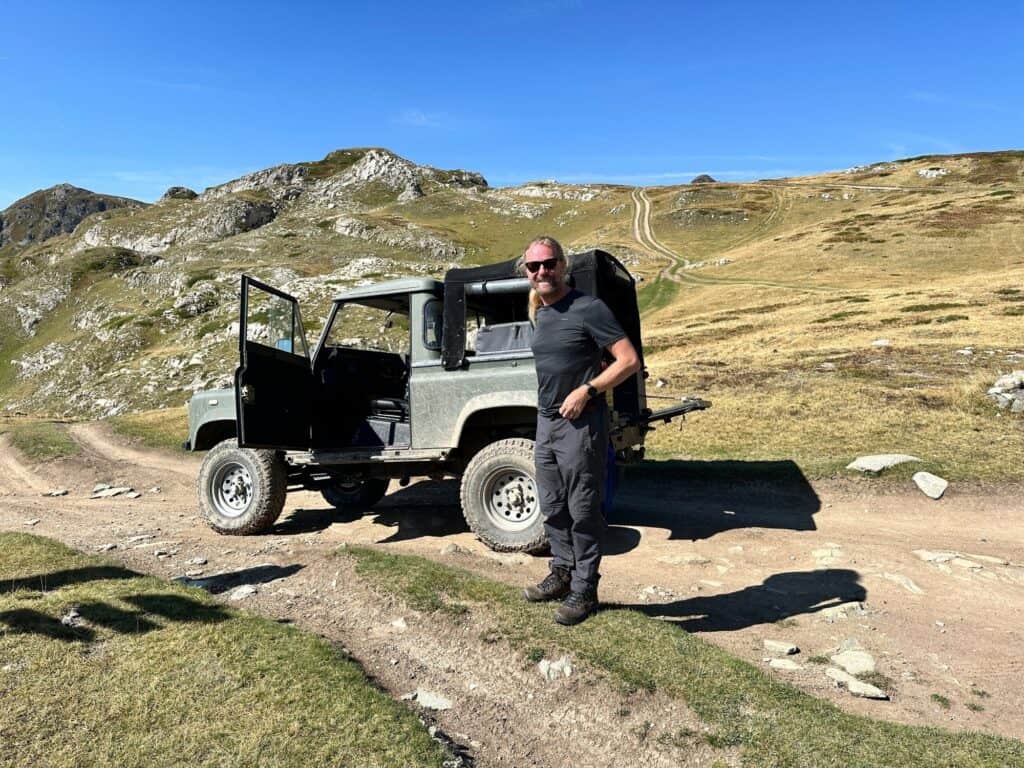 man standng in front of an old jeep on a dirt track - surrounded by high mountains 